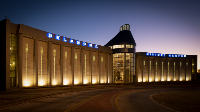 The Oklahoma History Center building in the evening, with glowing light visible from the windows and signage.