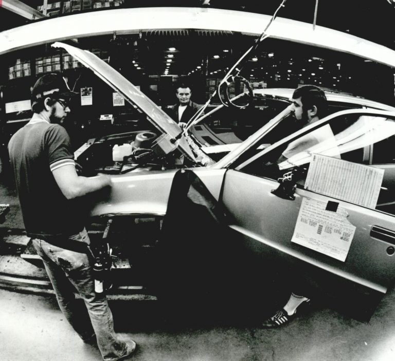 Historic photograph of three men standing around a car with its hood open inside the G M plant
