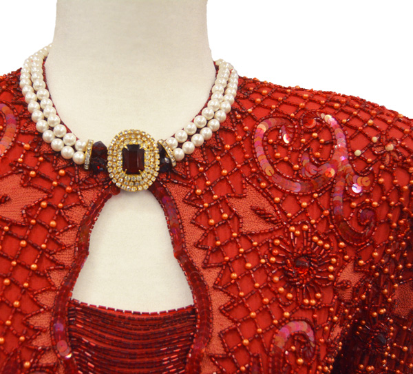 Close-up of the top of a red ball gown with intricate red beads of various sizes, and a necklace of pearls and red stones.