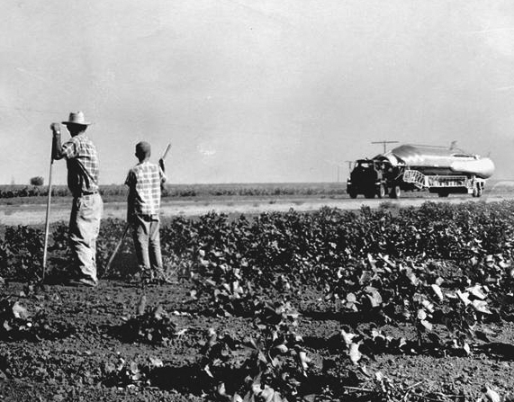 A man and a boy in a field hold farming implements as they watch a large truck hauling a missile.
